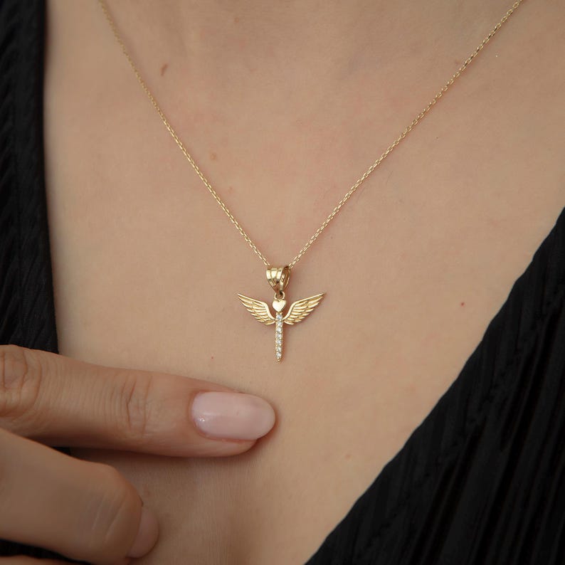May include: A gold necklace with a pendant shaped like an angel with wings and a heart in the center. The angel is adorned with small clear stones.