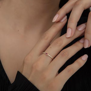 Könnte beinhalten: Ein goldener Ring mit einem rechteckigen, roten Edelstein. Das Band ist mit kleinen, klaren Edelsteinen verziert. Der Ring wird auf einer Hand mit gepflegten Nägeln vor dunklem Hintergrund gezeigt.