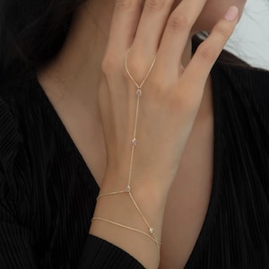 Könnte beinhalten: Eine zarte goldene Handkette mit kleinen, funkelnden Edelsteinen. Die Kette erstreckt sich vom Handgelenk über den Handrücken bis zum Mittelfinger. Der Schmuck wird vor einem schwarzen Hintergrund präsentiert.