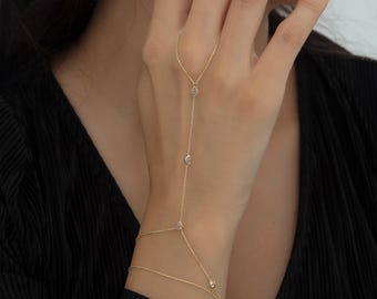 May include: A delicate gold hand chain with small, sparkling gemstones. The chain extends from the wrist, across the back of the hand, and up to the middle finger. The jewelry is displayed against a black background.