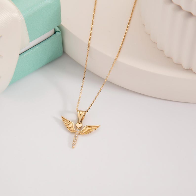 May include: A gold chain necklace with a small angel pendant featuring a heart with clear stones. The angel has wings and a heart in the center.