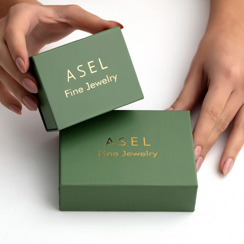 May include: Two sage green jewelry boxes with gold lettering that reads "ASEL Fine Jewelry". The boxes are rectangular and have a matte finish. One box is open, revealing the inside, while the other is closed.
