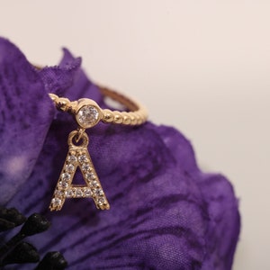 Könnte beinhalten: Ein goldener Ring mit einem kleinen, klaren Stein und einem Anhänger in Form des Buchstabens 'A', der mit klaren Steinen besetzt ist. Der Ring liegt auf einem violetten Stoffhintergrund.