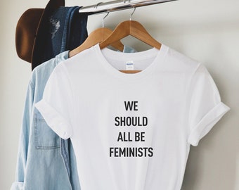 May include: White t-shirt with the text "WE SHOULD ALL BE FEMINISTS" printed in black. The shirt is on a hanger and has a knotted bottom.