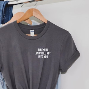 Puede incluir: Camiseta gris con el texto "Bisexual y aún no interesado en ti" en letras blancas. La camiseta está colgada en una percha de madera.