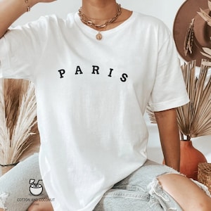 May include: White oversized t-shirt with the word "PARIS" printed in black on the front. The shirt is being worn by a person with a gold necklace.