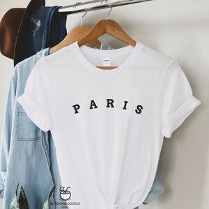 May include: White t-shirt with black text that reads "PARIS" on a hanger. The shirt is folded and tied at the bottom.