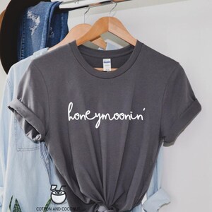 May include: A gray t-shirt with the text "honeymoonin'" in white lettering. The shirt is hanging on a wooden hanger.