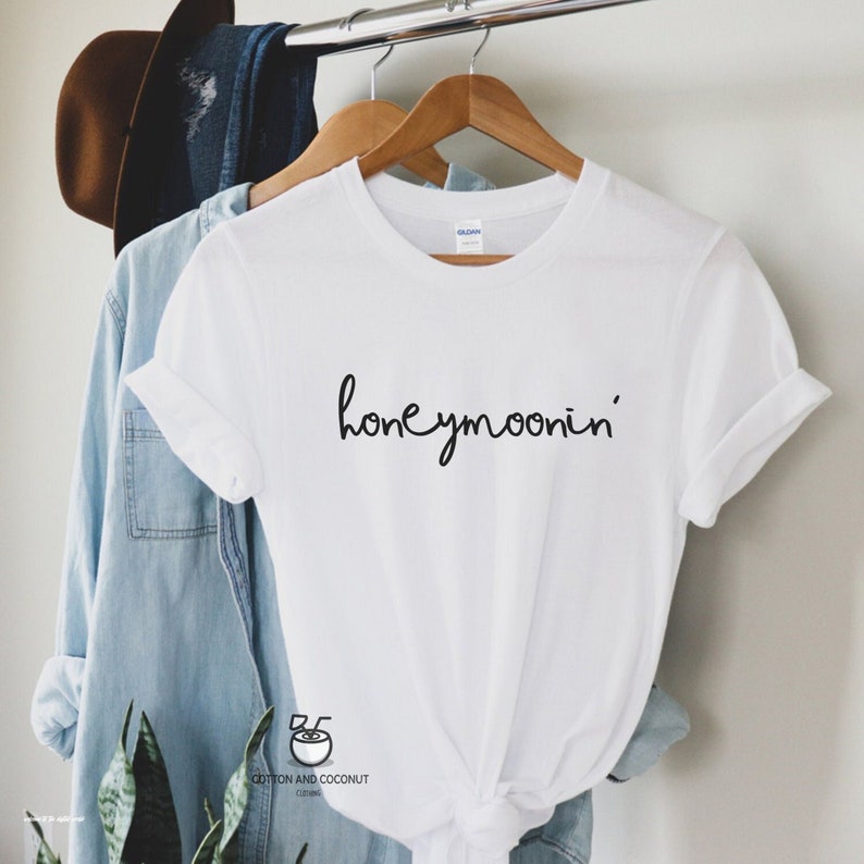 May include: White t-shirt with black text that says "honeymoonin'" on a hanger. The shirt is tied at the bottom.
