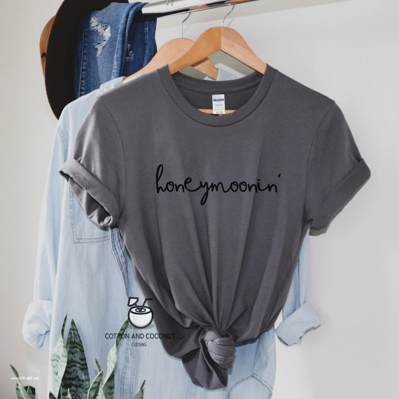 May include: A gray t-shirt with the text "honeymoonin'" in black script on the front. The shirt is hanging on a wooden hanger.