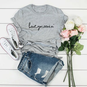 May include: Gray t-shirt with the text "honeymoonin'" in black script. The shirt is paired with denim shorts and white sneakers.