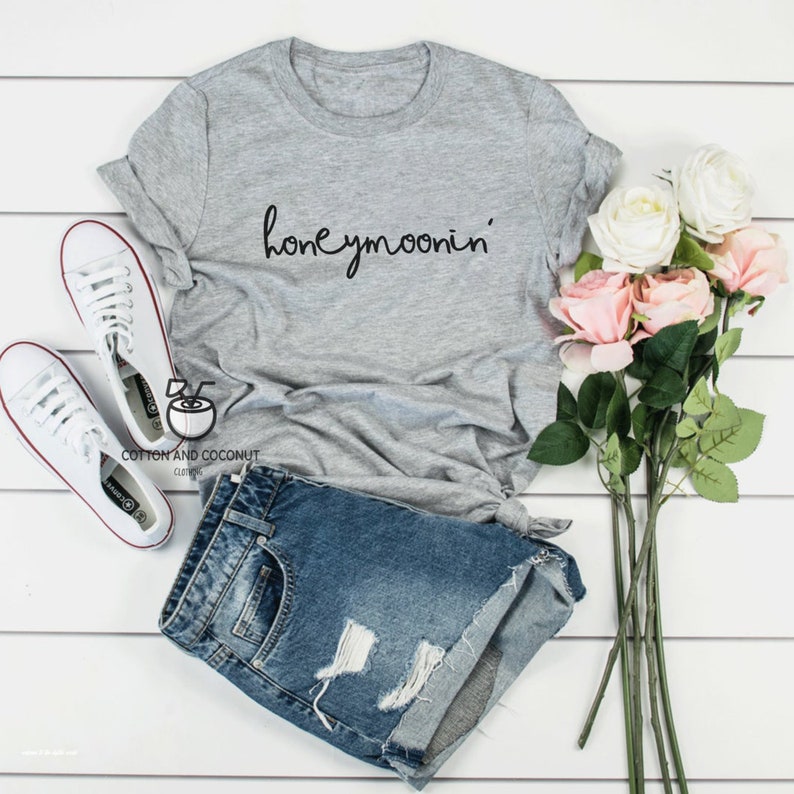 May include: Gray t-shirt with the text "honeymoonin'" in black script. The shirt is paired with denim shorts and white sneakers.