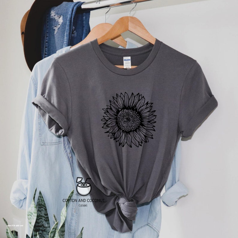 Puede incluir: Una camiseta gris antracita con un gr&aacute;fico de girasol en blanco y negro. La camiseta est&aacute; en una percha y tiene cuello redondo.