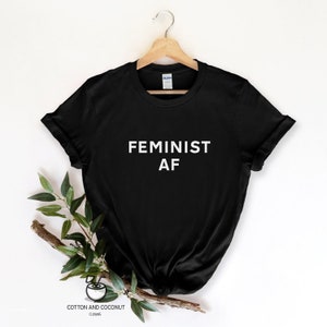 Puede incluir: Camiseta negra con el texto blanco "FEMINIST AF" mostrado. La camiseta est&aacute; colgada de una percha de madera sobre un fondo blanco, con una ramita de vegetaci&oacute;n a un lado. La marca "Cotton and Coconut Clothing" est&aacute; impresa en la parte inferior.
