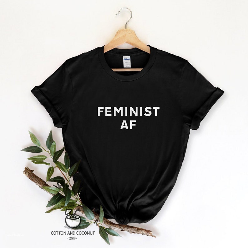 Puede incluir: Camiseta negra con el texto blanco "FEMINIST AF" mostrado. La camiseta est&aacute; colgada de una percha de madera sobre un fondo blanco, con una ramita de vegetaci&oacute;n a un lado. La marca "Cotton and Coconut Clothing" est&aacute; impresa en la parte inferior.