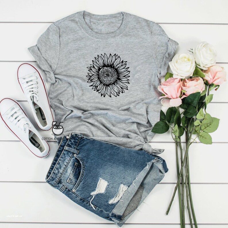 Puede incluir: Una camiseta gris con un gr&aacute;fico de girasol en blanco y negro. La camiseta se combina con pantalones cortos de mezclilla y zapatillas blancas.