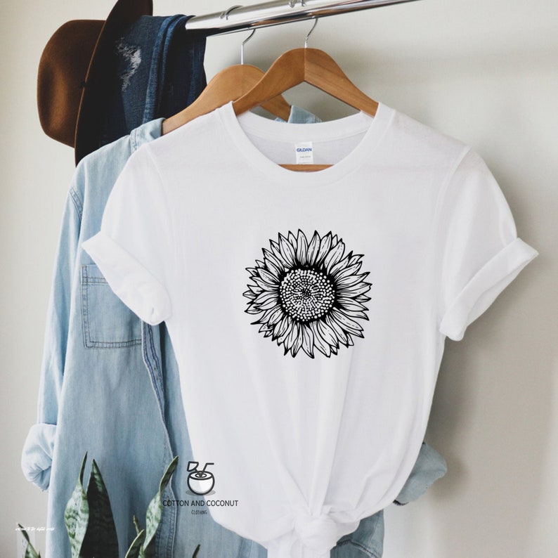 Puede incluir: Camiseta blanca con un dibujo en blanco y negro de un girasol. La camiseta est&aacute; en una percha y tiene el texto "Cotton and Coconut Clothing" en la parte inferior.