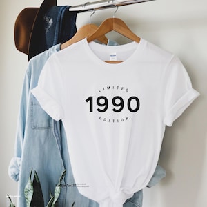 Puede incluir: Camiseta blanca con el texto "LIMITED 1990 EDITION" en un diseño circular negro. La camiseta está colgada en una percha de madera, con una camisa vaquera y un sombrero marrón en el fondo.