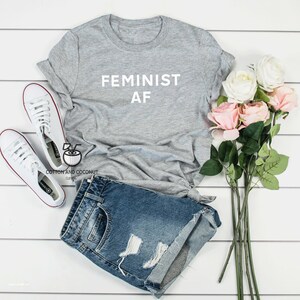 Puede incluir: Una camiseta gris con el texto blanco "FEMINIST AF", combinada con pantalones cortos de mezclilla y zapatillas blancas. Un ramo de rosas rosas y blancas a&ntilde;ade un toque de color al atuendo informal, sobre un fondo de madera blanca.