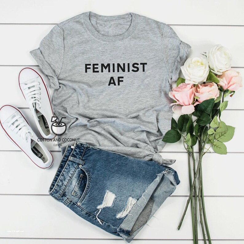 Puede incluir: Una camiseta gris con la frase "FEMINIST AF" en letras negras. Se combina con un pantal&oacute;n corto vaquero, zapatillas blancas y un ramo de rosas rosas y blancas. La imagen est&aacute; dispuesta sobre una superficie de madera blanca.