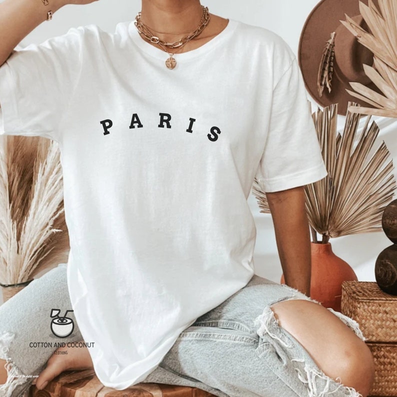 May include: White oversized t-shirt with the word "PARIS" printed in black on the front. The shirt is being worn by a person with a gold necklace.