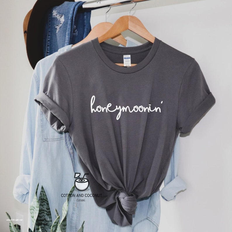 May include: A gray t-shirt with the text "honeymoonin'" in white lettering. The shirt is hanging on a wooden hanger.
