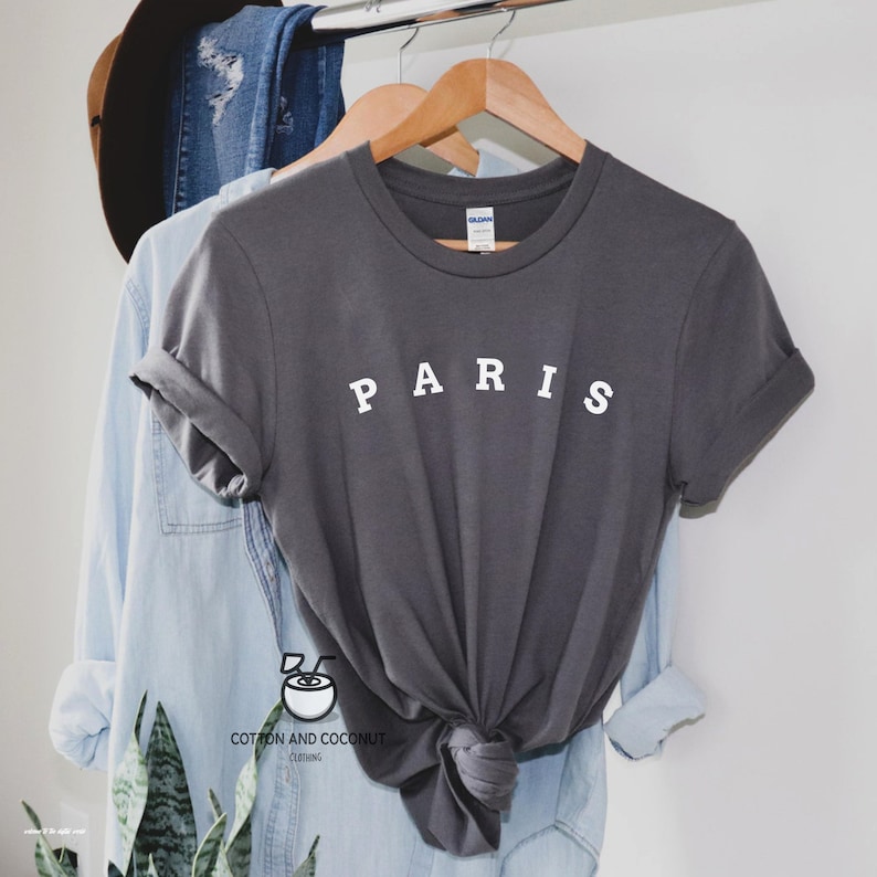 May include: A charcoal gray t-shirt with the word "PARIS" printed in white on the front. The shirt is folded and hanging on a wooden hanger.