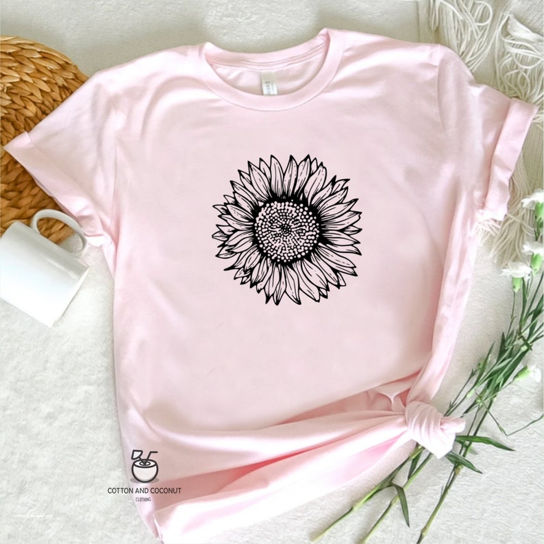 Puede incluir: Una camiseta rosa con un dibujo de l&iacute;nea negra de un girasol en la parte delantera. La camiseta est&aacute; hecha de un material suave y c&oacute;modo y tiene un cuello redondo.
