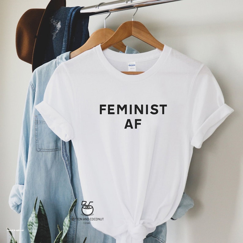Puede incluir: Camiseta blanca con el texto "FEMINIST AF" en negro, colgada de una percha de madera. Una camisa vaquera azul claro y un sombrero marr&oacute;n son visibles en el fondo. La camiseta es de Cotton and Coconut Clothing.