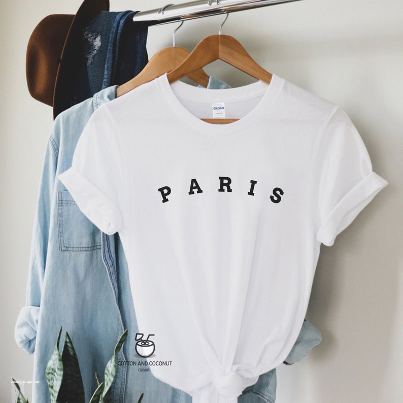 May include: White t-shirt with black text that reads "PARIS" on a hanger. The shirt is folded and tied at the bottom.