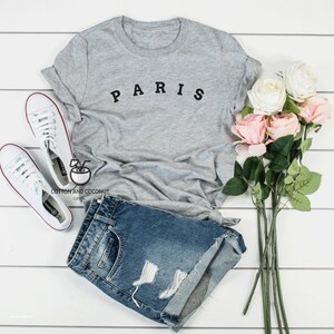 May include: A gray t-shirt with the word "PARIS" printed in black letters. The shirt is paired with denim shorts and white sneakers. A bouquet of pink and white roses is in the background.