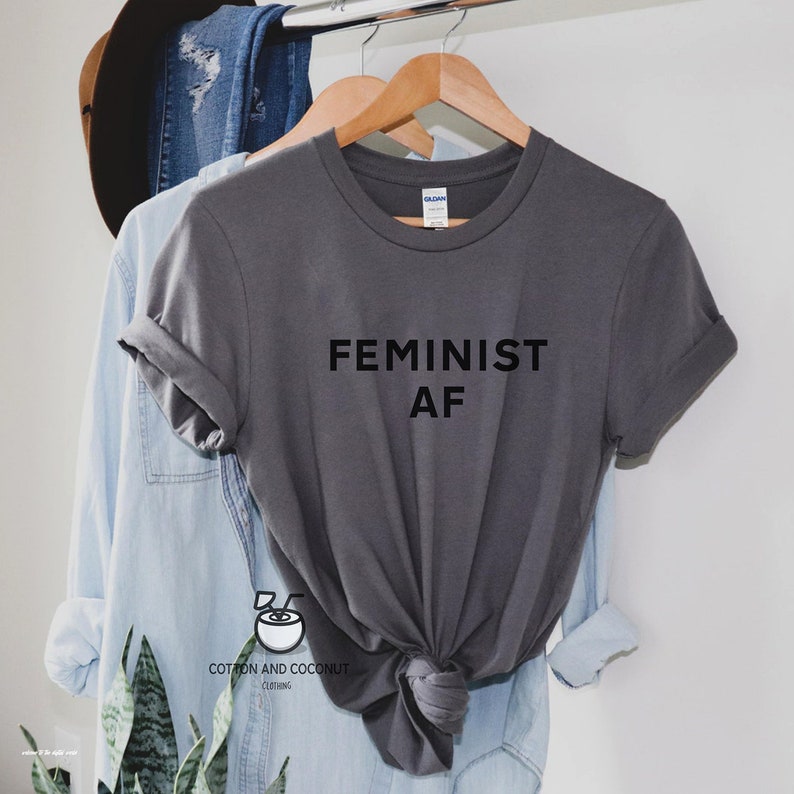 Puede incluir: Una camiseta gris con las palabras "FEMINIST AF" impresas en negro. La camiseta est&aacute; colgada en una percha de madera, con una camisa abotonada azul claro y unos vaqueros azules en el fondo. La marca "Cotton and Coconut Clothing" est&aacute; impresa en la parte inferior.