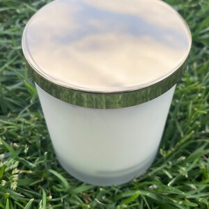 K&ouml;nnte beinhalten: Ein wei&szlig;es Kerzenglas aus mattiertem Glas mit einem goldfarbenen Deckel und Rand. Die Kerze steht auf einem Bett aus gr&uuml;nem Gras. Der Deckel reflektiert den Himmel.