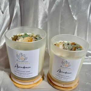 K&ouml;nnte beinhalten: Zwei kristallinfundierte Sojakerzen in Gl&auml;sern aus mattiertem Glas. Jede Kerze hat ein wei&szlig;es Etikett mit dem Text "Abundance" und "Eva Luna Crystals". Die Kerzen sind mit Kristallen bedeckt und stehen auf Holzuntersetzern.