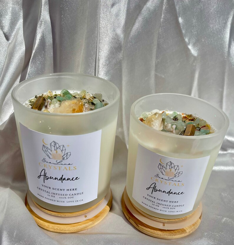 K&ouml;nnte beinhalten: Zwei kristallinfundierte Sojakerzen in Gl&auml;sern aus mattiertem Glas. Jede Kerze hat ein wei&szlig;es Etikett mit dem Text "Abundance" und "Eva Luna Crystals". Die Kerzen sind mit Kristallen bedeckt und stehen auf Holzuntersetzern.