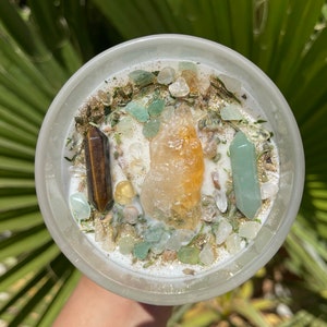 K&ouml;nnte beinhalten: Eine wei&szlig;e Kerze in einem transparenten Glasbeh&auml;lter, verziert mit Kristallen und Kr&auml;utern. Die Kerze hat einen gro&szlig;en orangefarbenen Kristall in der Mitte, flankiert von einem braunen und einem gr&uuml;nen Kristall. Gr&uuml;ne und klare Steine ​​und goldener Glitzer sind verstreut.
