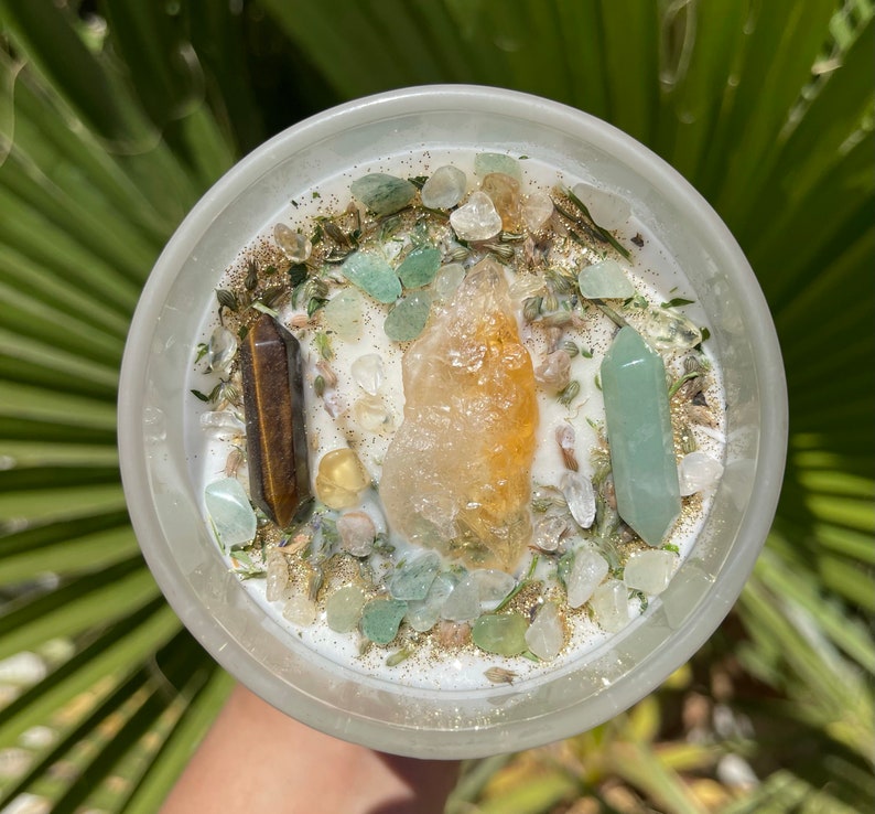 K&ouml;nnte beinhalten: Eine wei&szlig;e Kerze in einem transparenten Glasbeh&auml;lter, verziert mit Kristallen und Kr&auml;utern. Die Kerze hat einen gro&szlig;en orangefarbenen Kristall in der Mitte, flankiert von einem braunen und einem gr&uuml;nen Kristall. Gr&uuml;ne und klare Steine ​​und goldener Glitzer sind verstreut.