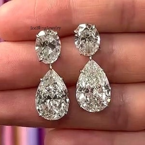 May include: A pair of diamond drop earrings. Each earring features a sparkling oval-cut diamond at the top, connected to a pear-shaped diamond below. The earrings are held up by a person's hand.