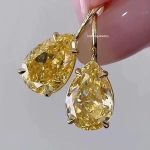 May include: A pair of drop earrings featuring large, pear-shaped yellow gemstones set in gold metal prongs with curved wire hooks. The text IceBlingJewelry is visible near the top of the setting. The faceted stones exhibit a bright yellow hue.
