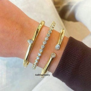 Könnte beinhalten: Vier goldfarbene Armbänder. Zwei sind massiv mit tropfenförmigen, klaren Steinen. Eines hat eine Reihe klarer Steine. Das vierte Armband hat an jedem Ende einen klaren Stein. Die Armbänder befinden sich an einem Handgelenk.