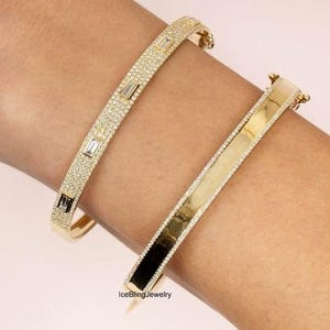 Può includere: Due braccialetti d'oro. Uno è incrostato di piccole pietre scintillanti e presenta accenti rettangolari. L'altro è una fascia d'oro lucida con una fila di piccole pietre lungo i bordi. I braccialetti sono sul braccio di una persona.