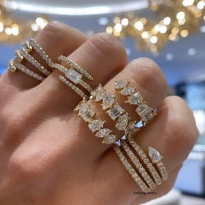 Può includere: Una mano che mostra diversi anelli d'oro con varie forme di diamanti. Gli anelli presentano diamanti taglio smeraldo, a goccia e rotondi, incastonati in sottili fasce. La disposizione mette in risalto i diversi design e la brillantezza.