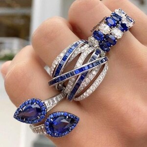 May include: A set of three rings featuring blue sapphire and white diamond stones. The rings are stacked on a finger. The top ring is a band of oval-shaped blue sapphires and white diamonds. The middle ring is a multi-band ring with alternating rows of blue sapphires and white diamonds. The bottom ring features two pear-shaped blue sapphires.