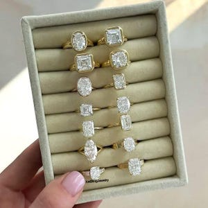 Könnte beinhalten: Eine Auswahl von 10 goldenen Verlobungsringen mit verschiedenen Diamantformen und -größen. Die Ringe sind in einer Reihe in einer weißen Samtbox angeordnet. Die Diamanten sind alle klar und funkeln.