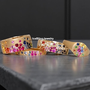 May include: Four gold-toned rings with colorful gemstones. The rings feature a variety of colored stones, including pink, blue, yellow, and orange. The rings are displayed on a dark gray surface.
