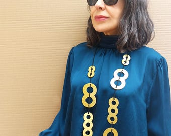 May include: A woman wearing a teal blue blouse with a gold and black geometric necklace. The necklace is made of large, round, interlocking circles.