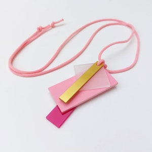 Könnte beinhalten: Eine geometrische Halskette in Rosa und Gold mit einer langen rosa Schnur. Der Anhänger besteht aus übereinanderliegenden rosa und klaren Acryl mit einer goldenen Stange.