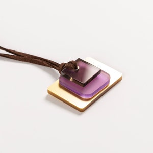 May include: A brown suede cord necklace with a layered pendant featuring a gold square and a smaller purple square.