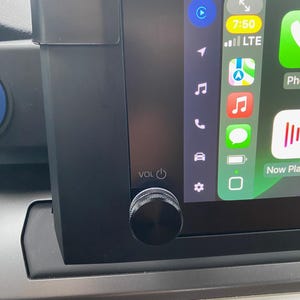 May include: A black car dashboard with a digital screen displaying a clock, music notes, and a phone icon. The screen also shows a notification for "Now Playing". A black volume knob with a power button is located below the screen.