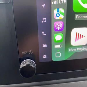 May include: A black knob with a power button labelled "VOL" is on a car dashboard. The dashboard screen shows a digital clock displaying 7:51, a signal strength indicator, the word "LTE", and several icons including a phone, music note, a car, and a gear.